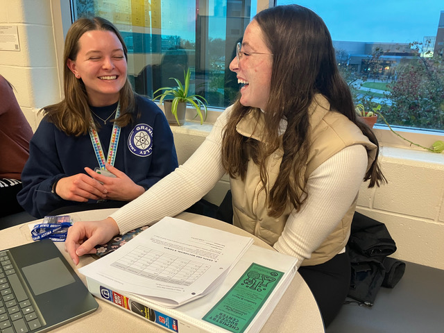 Two students laughing over an open binder of chemistry practice problems.